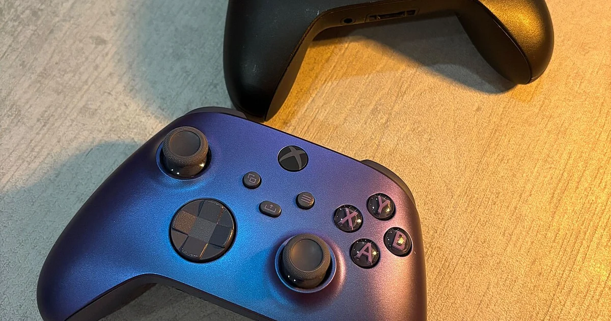 Two Xbox game controllers lie next to each other on a light, subtly textured surface. The top controller is black, while the bottom one has a blue-to-purple gradient finish. Both feature two analog sticks, a directional pad, menu and functi